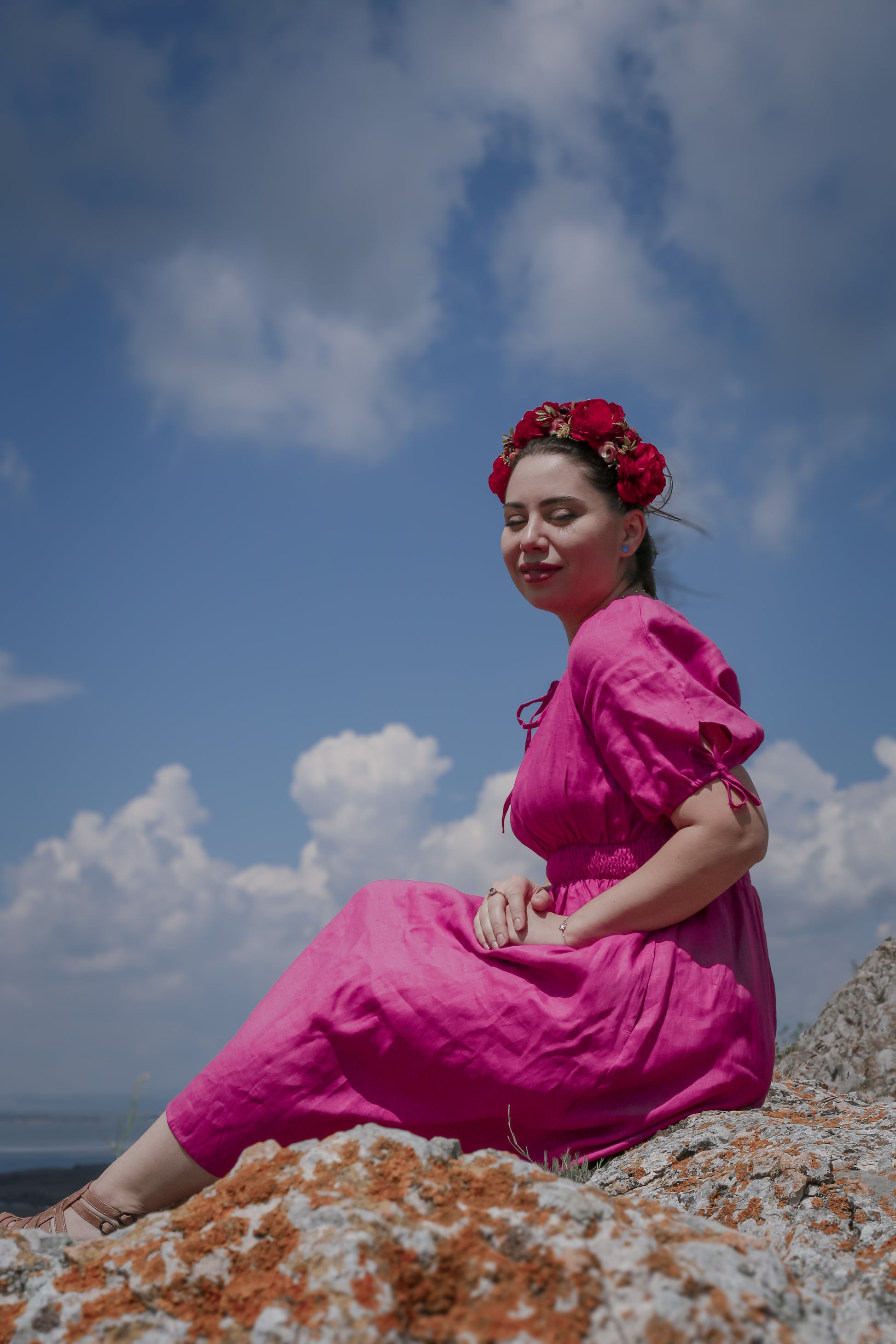 Woman in a pink modest nursing dress sitting on rocks with a blue sky and clouds in the background