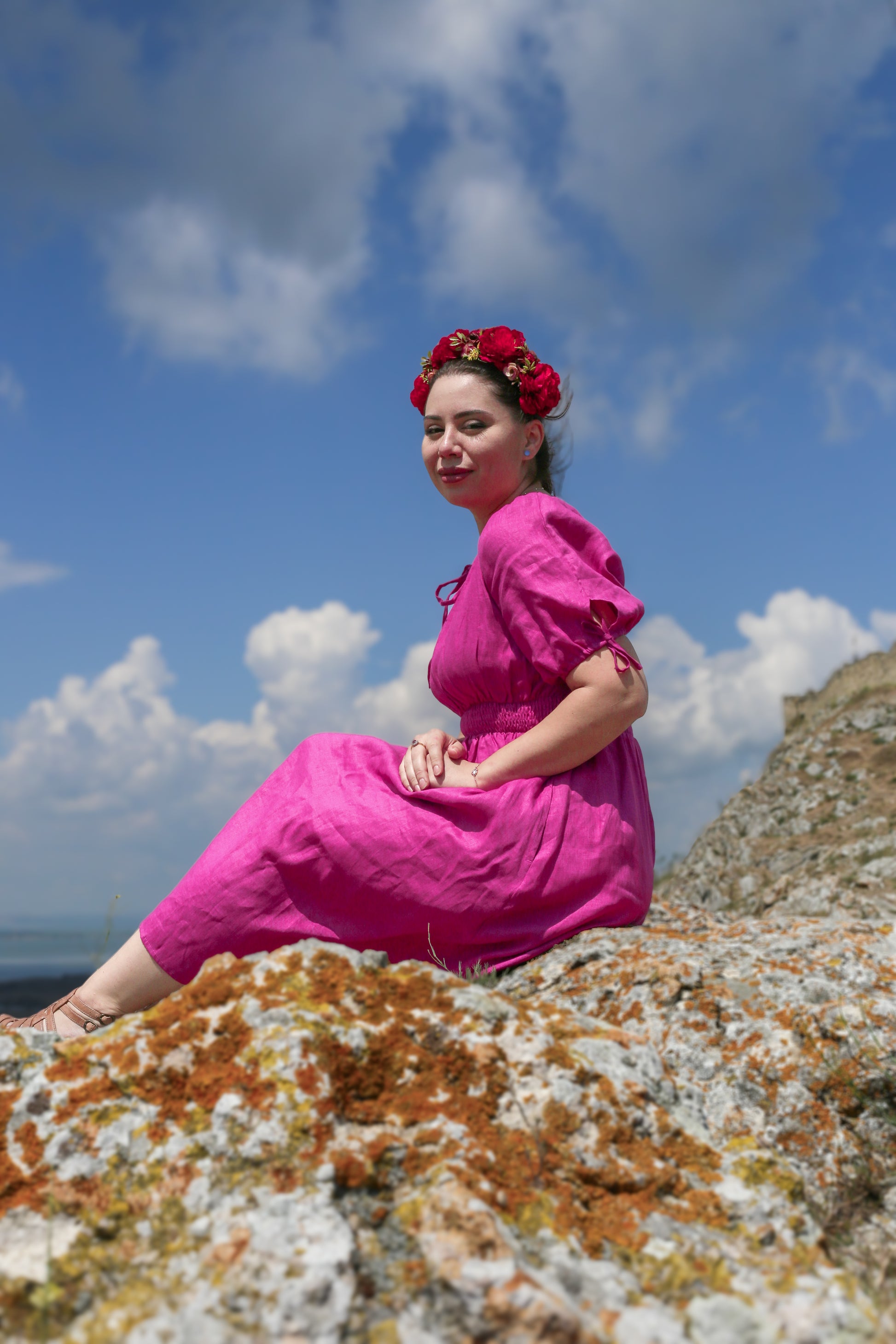 Woman in a pink modest nursing dress sitting on a rocky outcrop with a blue sky and clouds in the background.