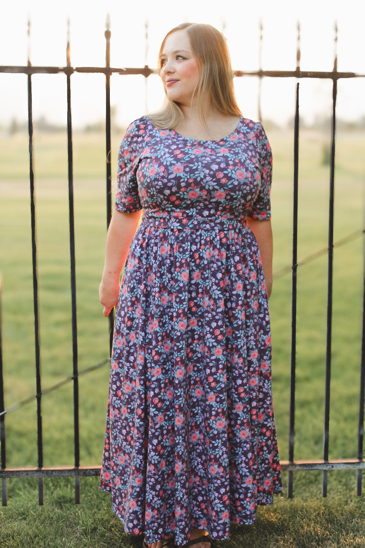Woman wearing a floral modest nursing dress standing in front of a metal fence with grass and sky in the background.