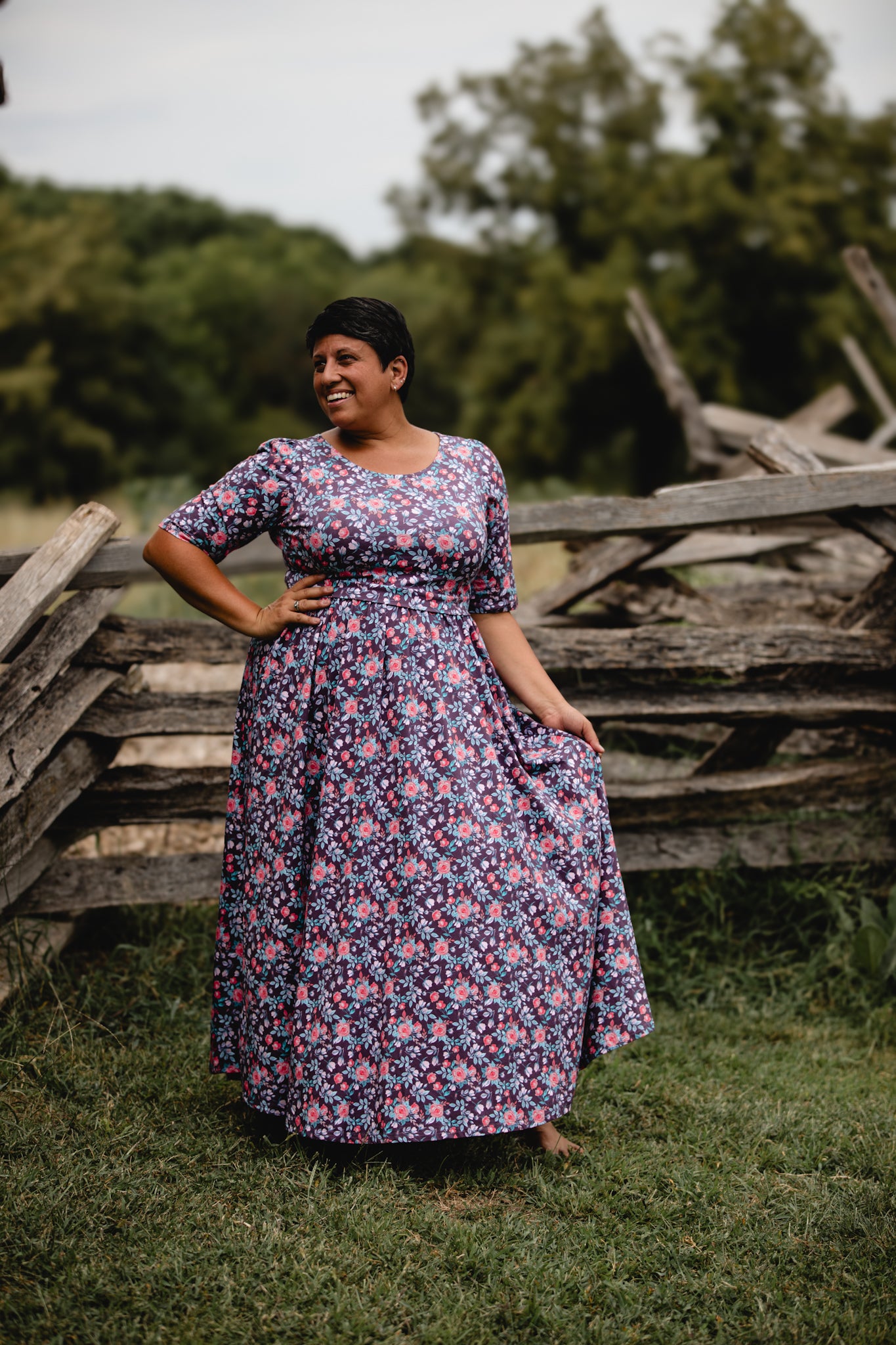 Woman in a modest nursing floral dress standing in a field with wooden fences and trees in the background