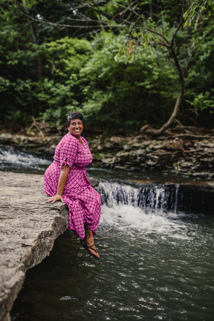 Woman in a pink modest nursing dress sitting on a rock by a small waterfall surrounded by greenery.