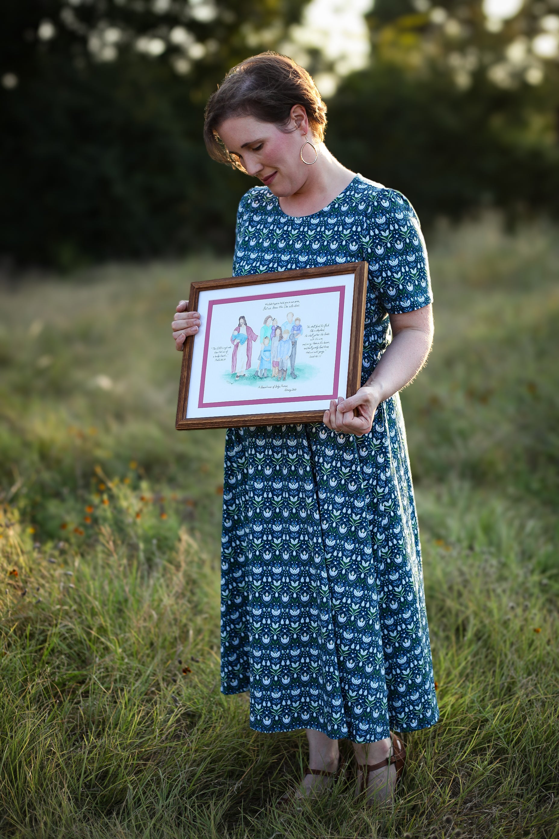 Woman in modest nursing dress with framed artwork, grassy field.