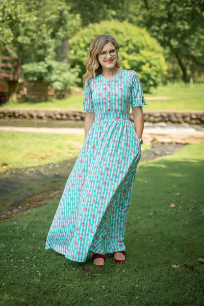 Woman in a long, patterned modest nursing dress standing outdoors on grass with trees in the background
