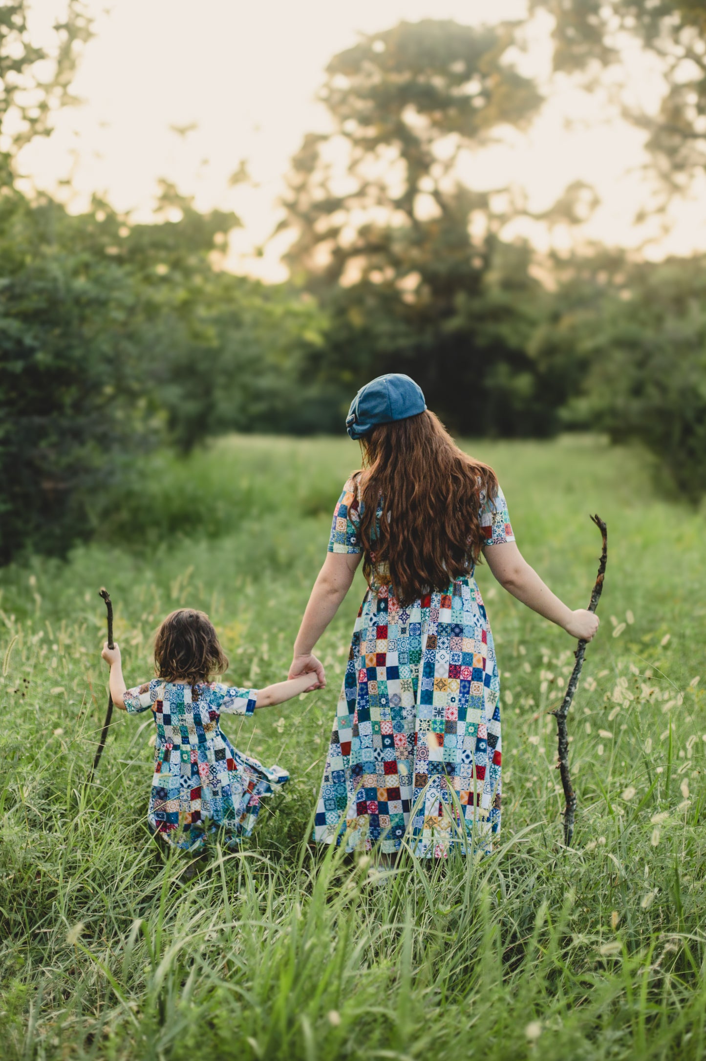 Mother and child in colorful modest dresses holding hands in a grassy field with trees in the background.