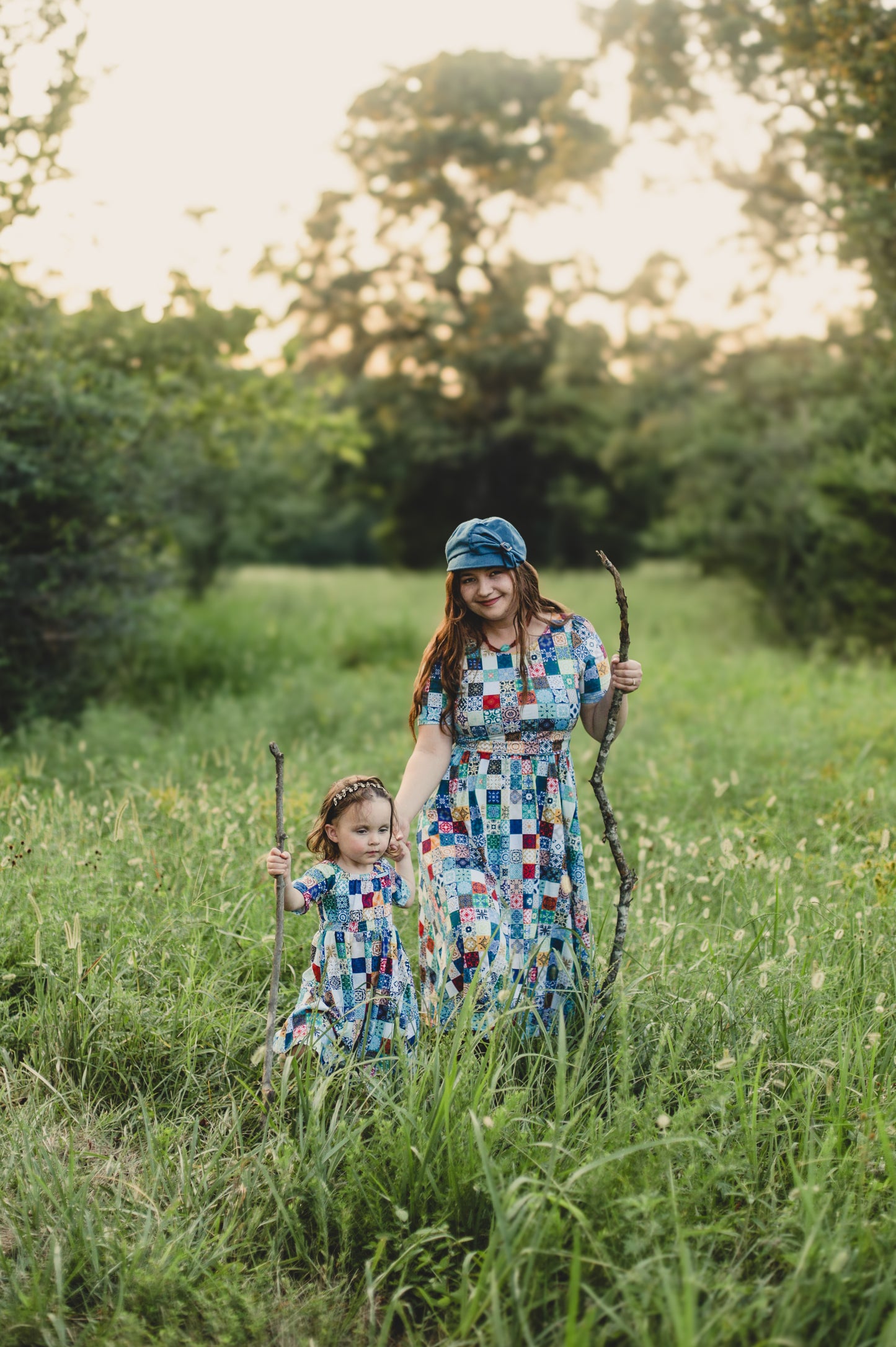 Mother and child in colorful checkered modest dresses holding sticks in a grassy field with trees in the background.