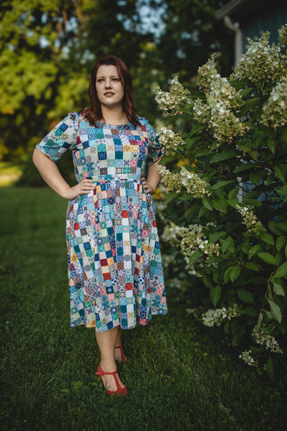 Woman wearing a colorful patchwork modest nursing dress standing outdoors near greenery.