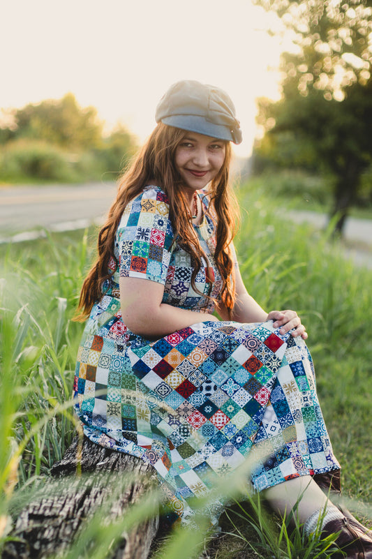 Woman sitting outdoors wearing a colorful patchwork modest nursing dress and cap.