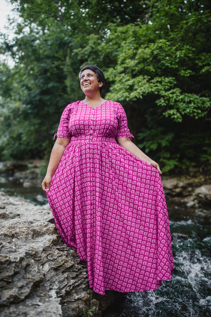 Woman in a pink modest nursing dress standing by a stream with trees in the background